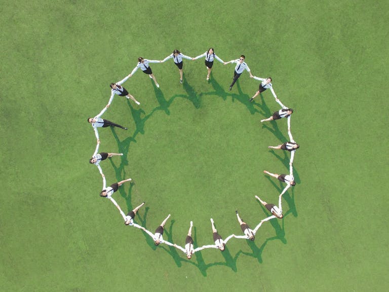 Aerial shot of a group forming a circle on a green field, holding hands and connected.