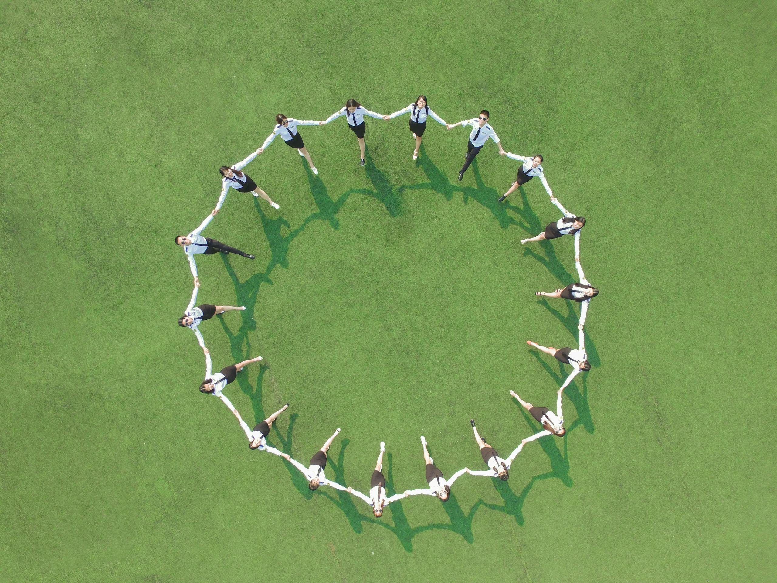 Aerial shot of a group forming a circle on a green field, holding hands and connected.