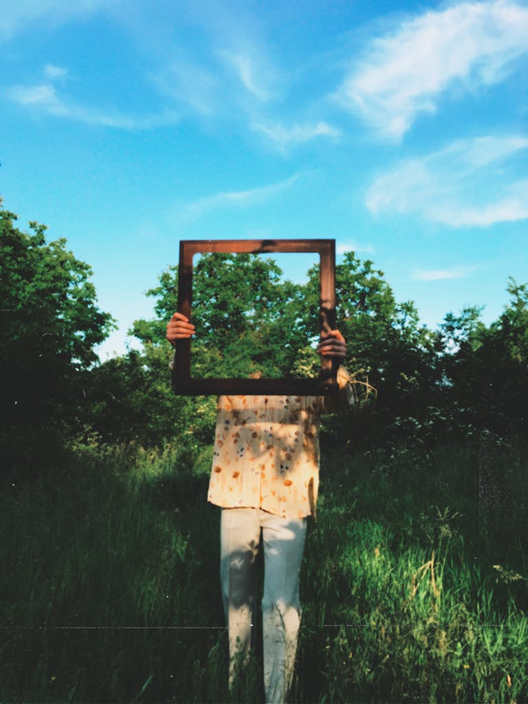 Creative portrait of an individual holding a frame in a vibrant green outdoor setting.