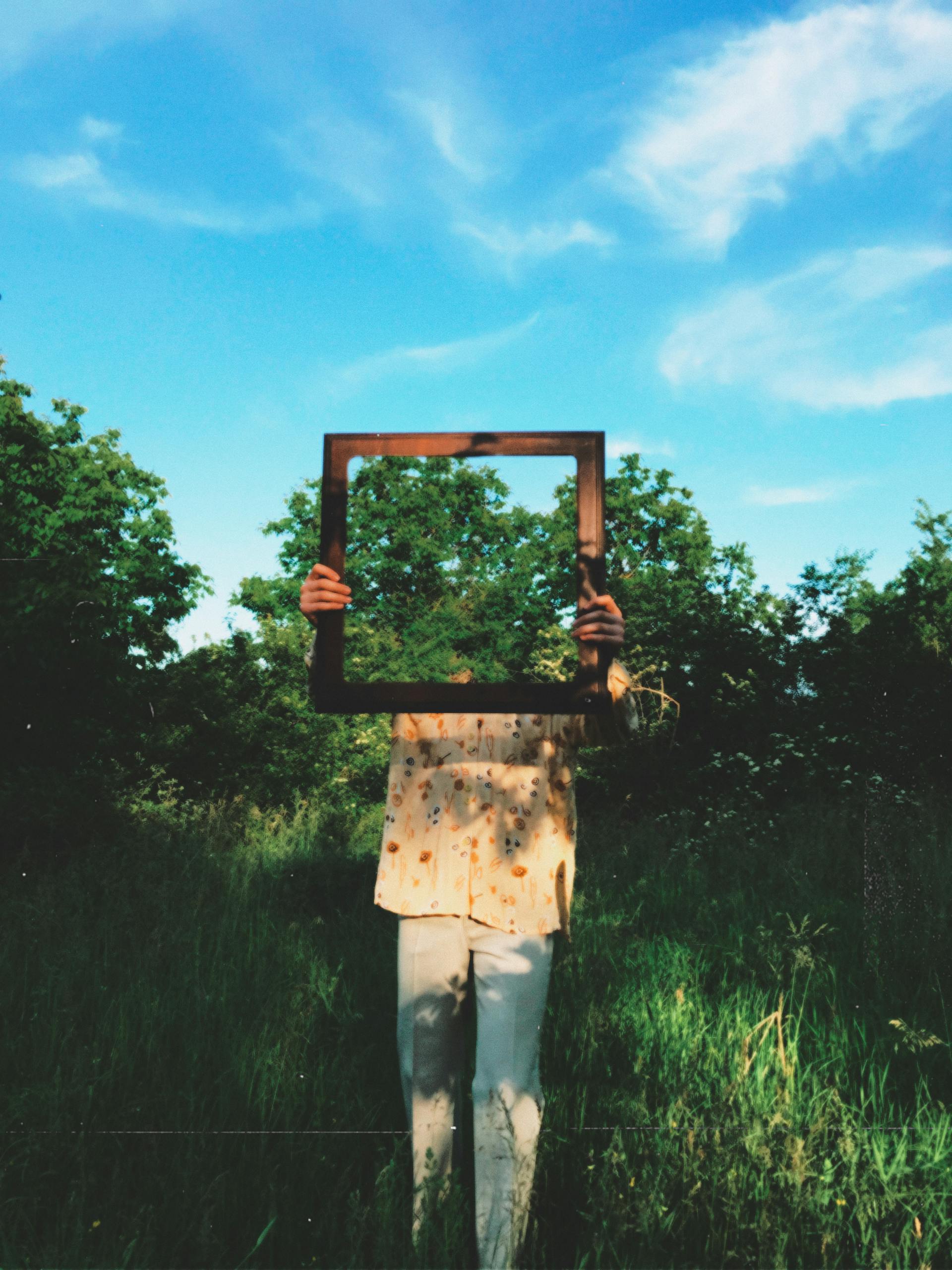 Creative portrait of an individual holding a frame in a vibrant green outdoor setting.